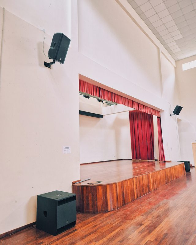 Hall stage with red curtain, wall-mounted loudspeakers and a floor-standing subwoofer